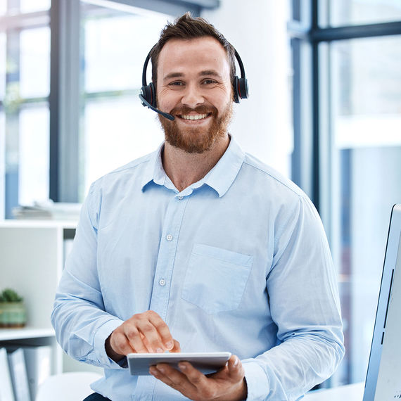 Jonge man met draadloze headset aan het werk op kantoor met tablet en vriendelijk glimlachend.